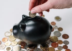 Hand inserting coin into a black piggy bank surrounded by various coins, symbolizing savings.